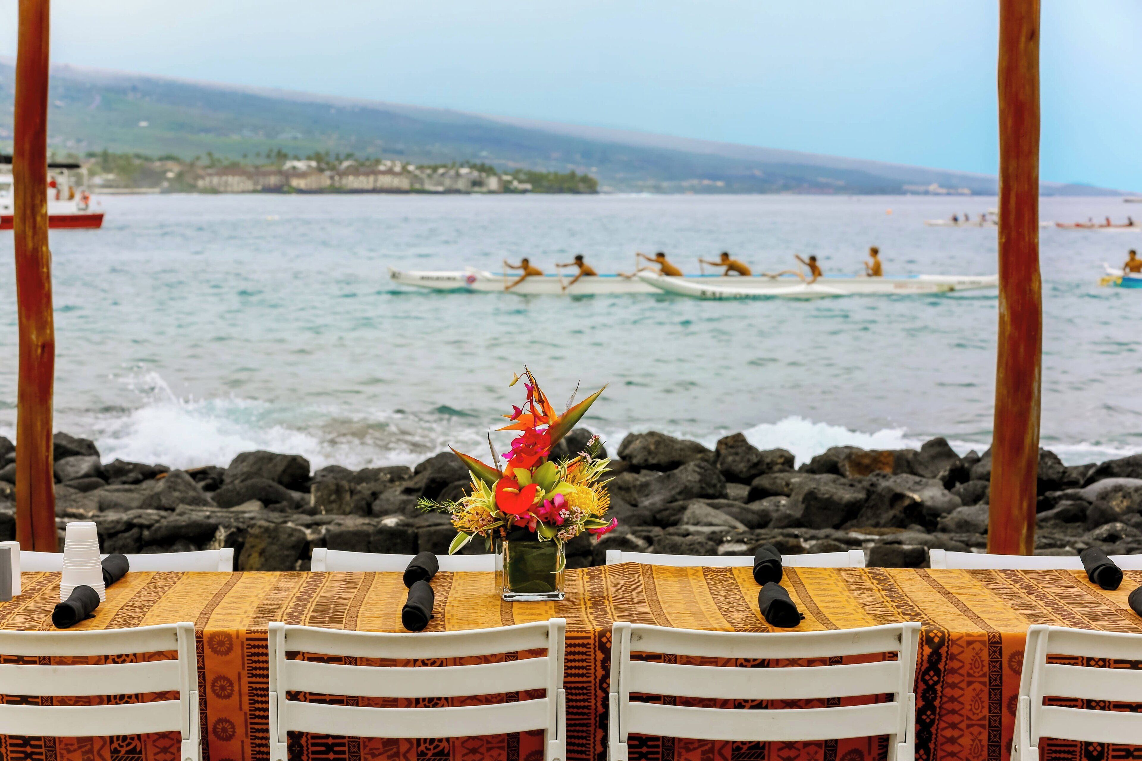Une table dressée pour le dîner, avec vue sur l’océan et une équipe d’aviron en arrière-plan.