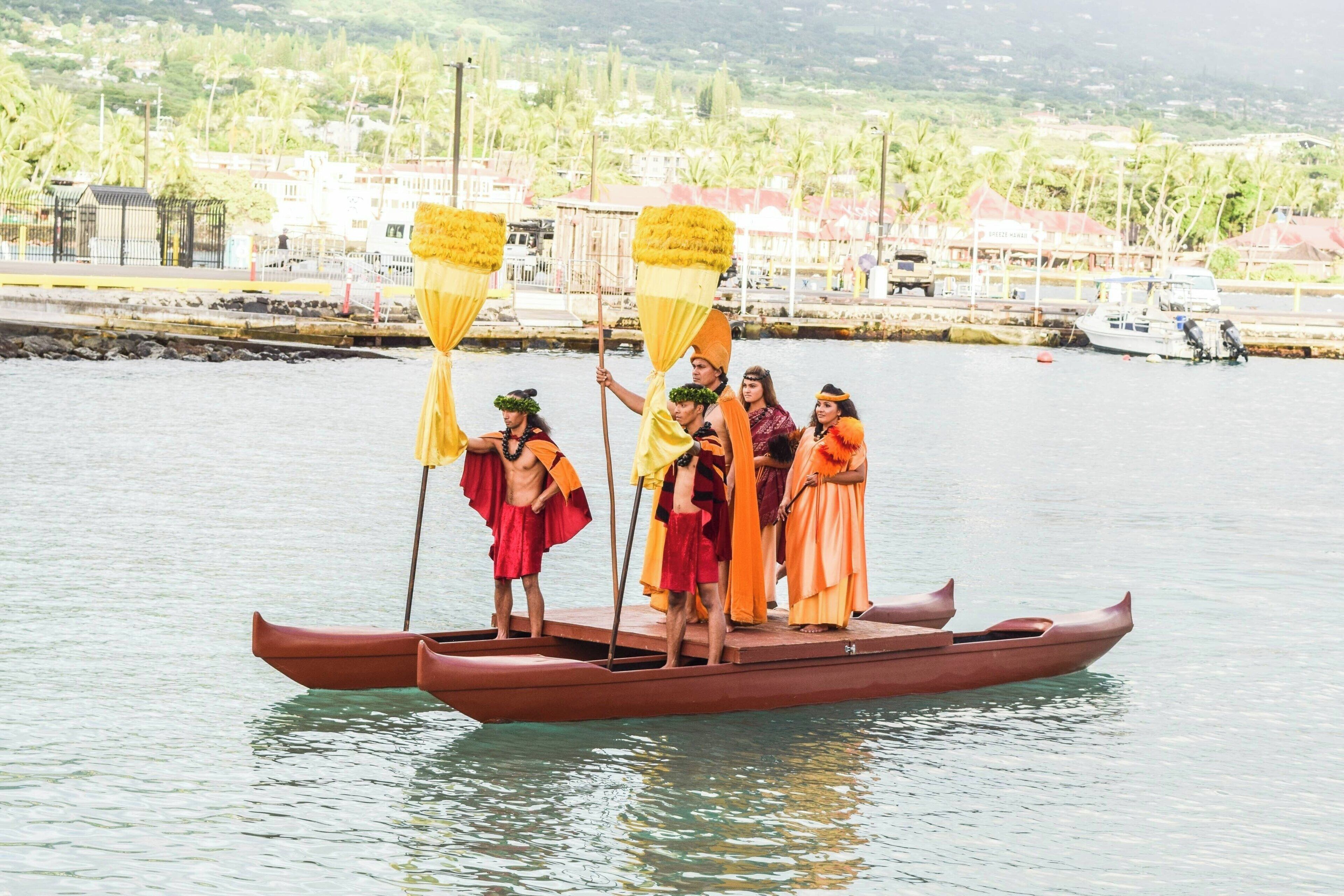 A group of people in traditional attire on a boat with a backdrop of a coastal town and greenery.