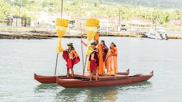 Un grupo de personas vestidas con atuendo tradicional en una embarcación, con un pueblo costero y vegetación de fondo.