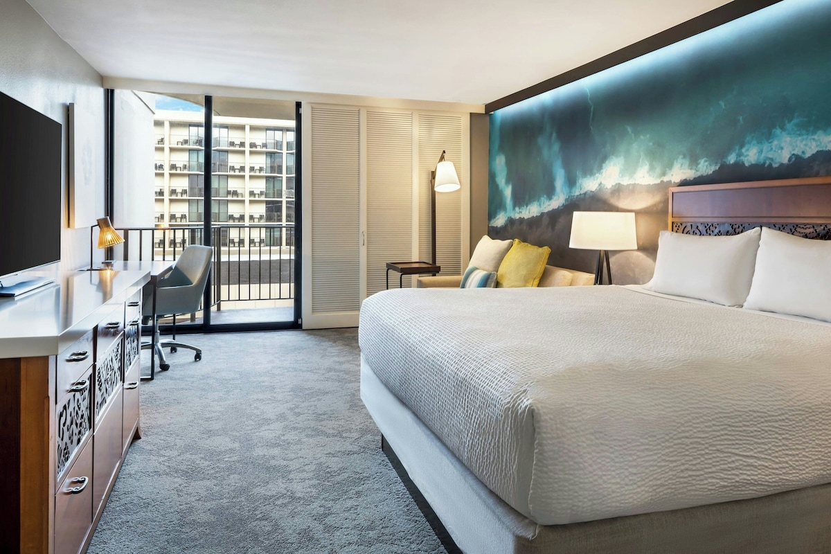 Ein Hotelzimmer mit einem großen Bett, einem Schreibtisch mit Stuhl, einem Fernseher und Blick auf ein Gebäude durch eine Glastür.