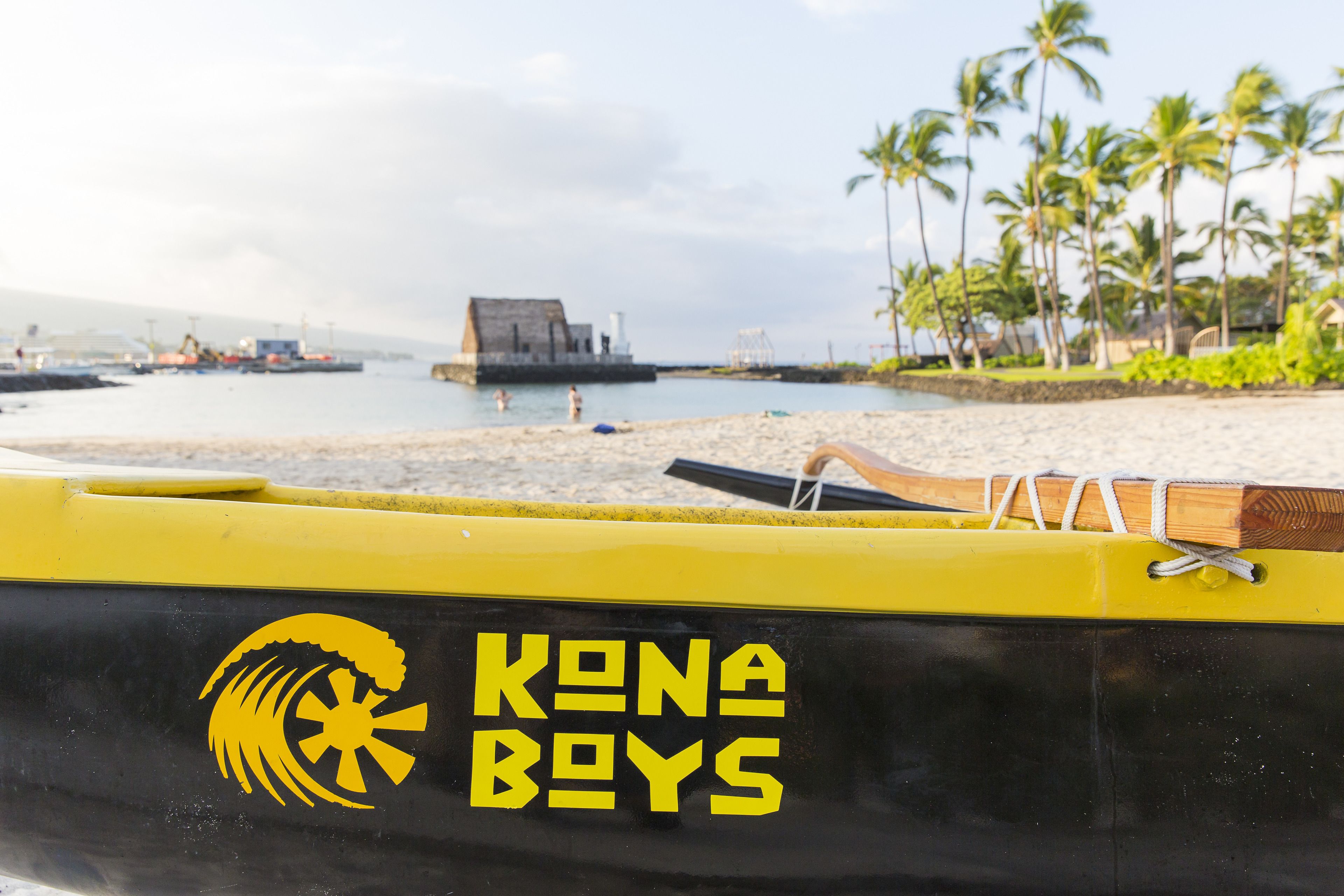 Eine Strandansicht mit einem gelben Boot, auf dem 'KONA BOYS' steht, und Palmen im Hintergrund.