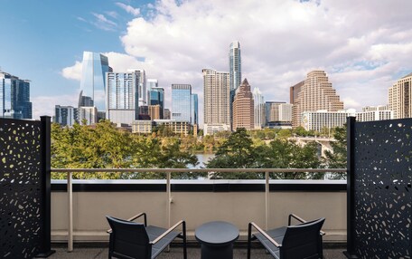 A balcony with two chairs and a table, overlooking a city skyline with modern buildings and a body of water.