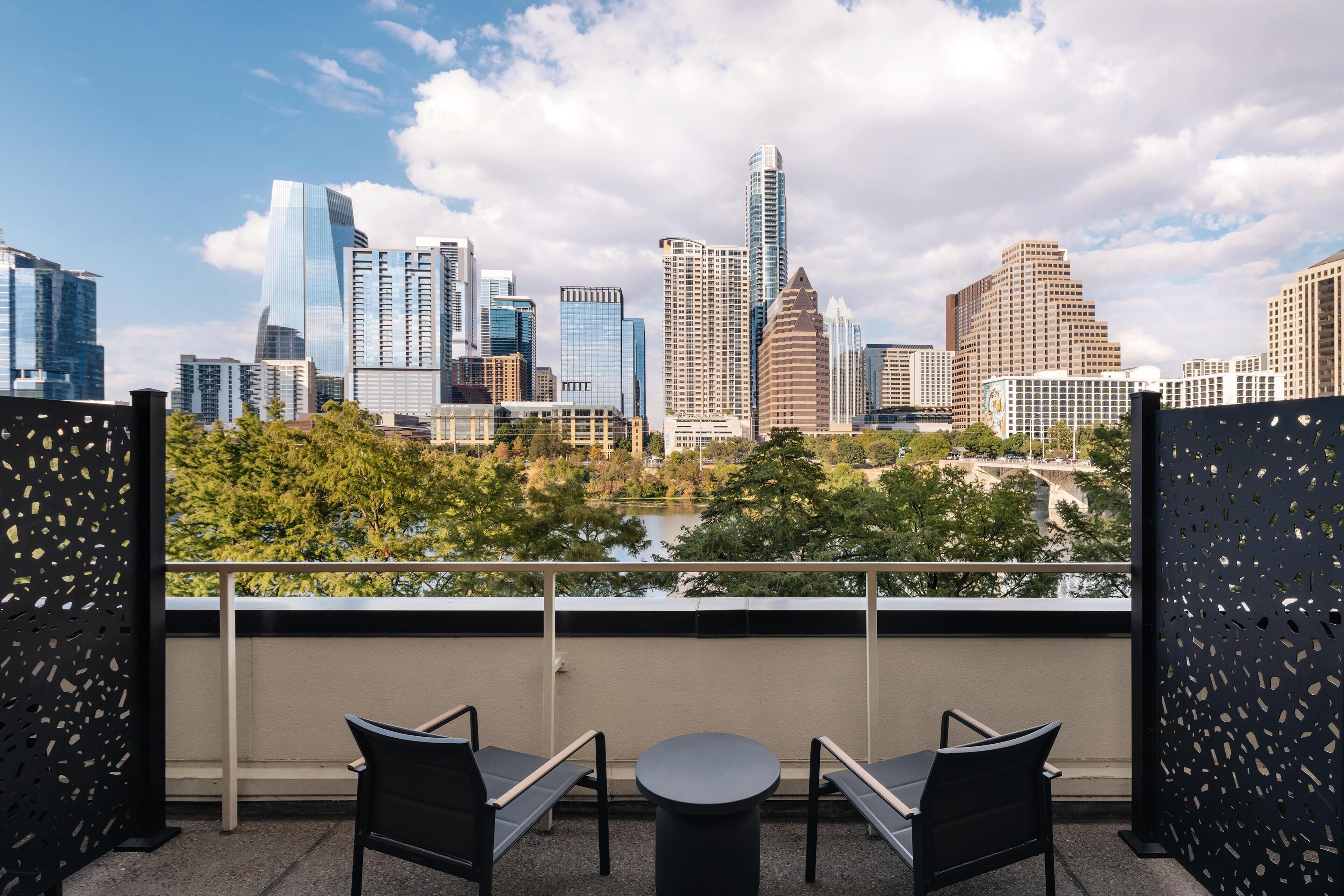 A balcony with two chairs and a table, overlooking a city skyline with various buildings and a body of water.