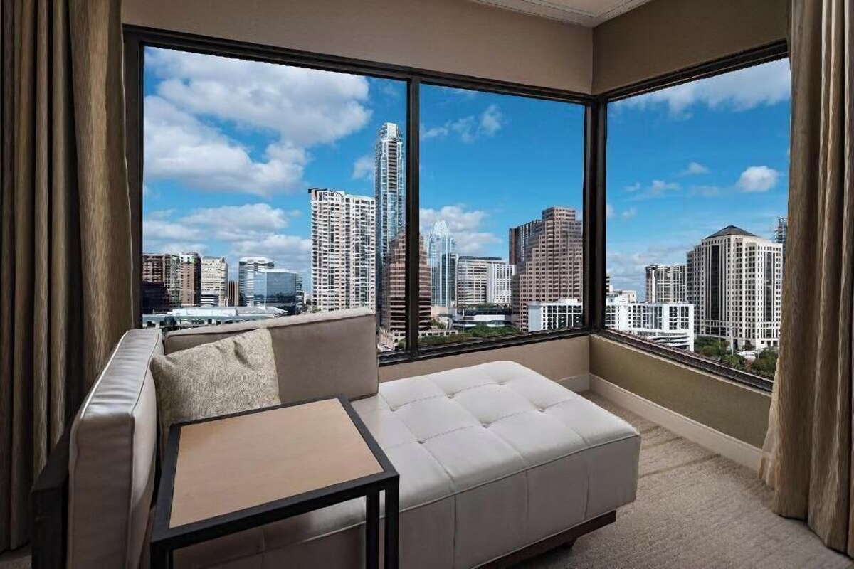 A modern hotel room with a large window offering a city skyline view, a white sofa, and a small table.