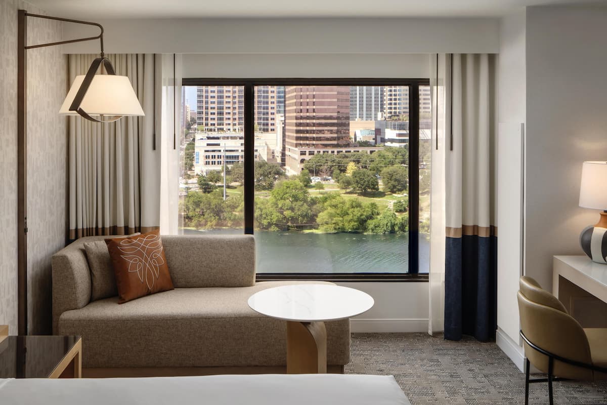 A hotel room with a sofa, a round table, a lamp, a view of a cityscape and a park through a large window.