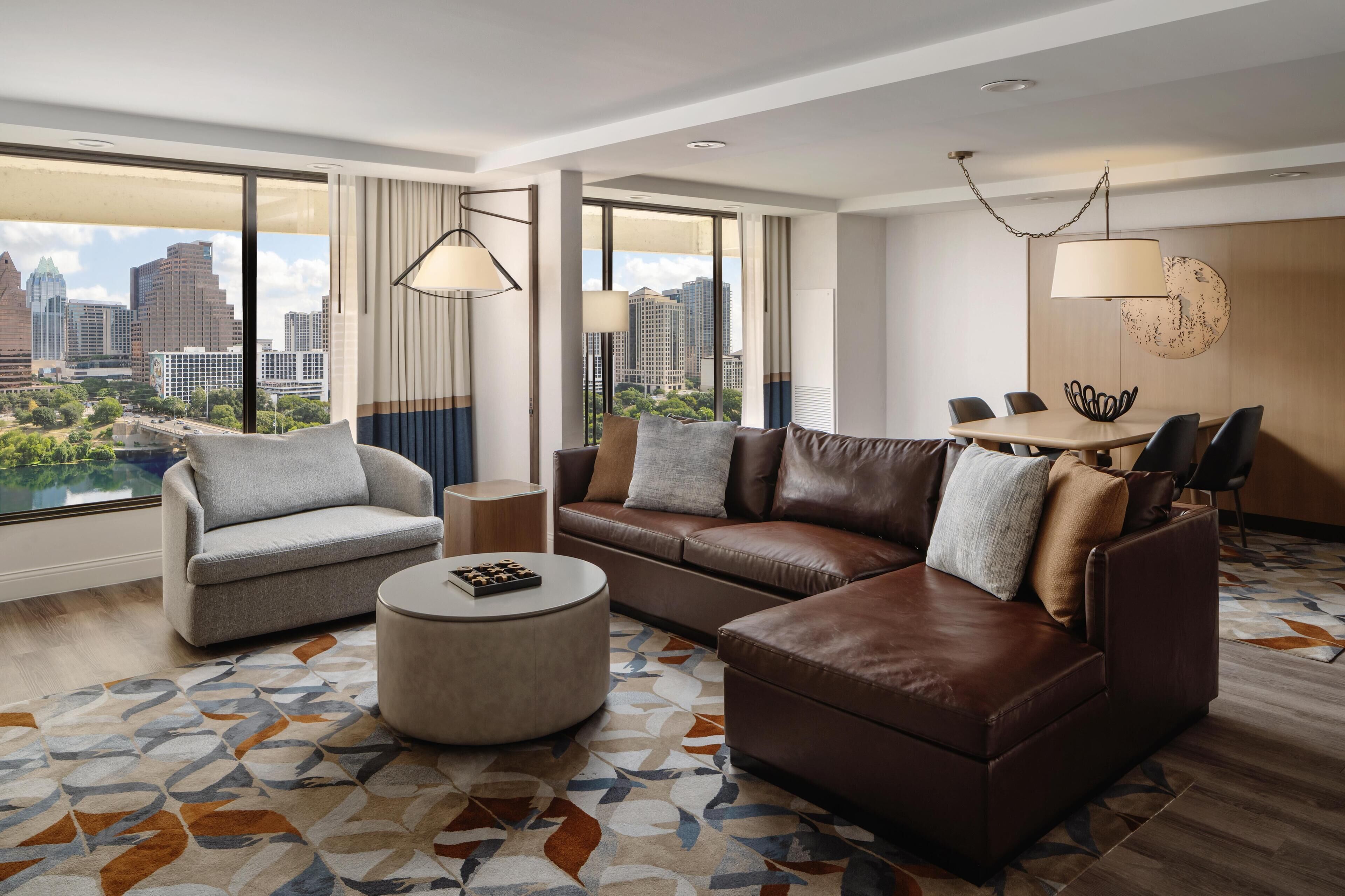 Een moderne hotelkamer met een groot raam, een bruine leren bank, een grijze fauteuil, een rond hockerkussen, en een zithoek met een tafel en stoelen.