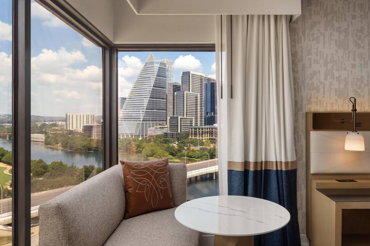 A hotel room with a large window, a sofa with a decorative pillow, a small round table, and a nightstand with a lamp.