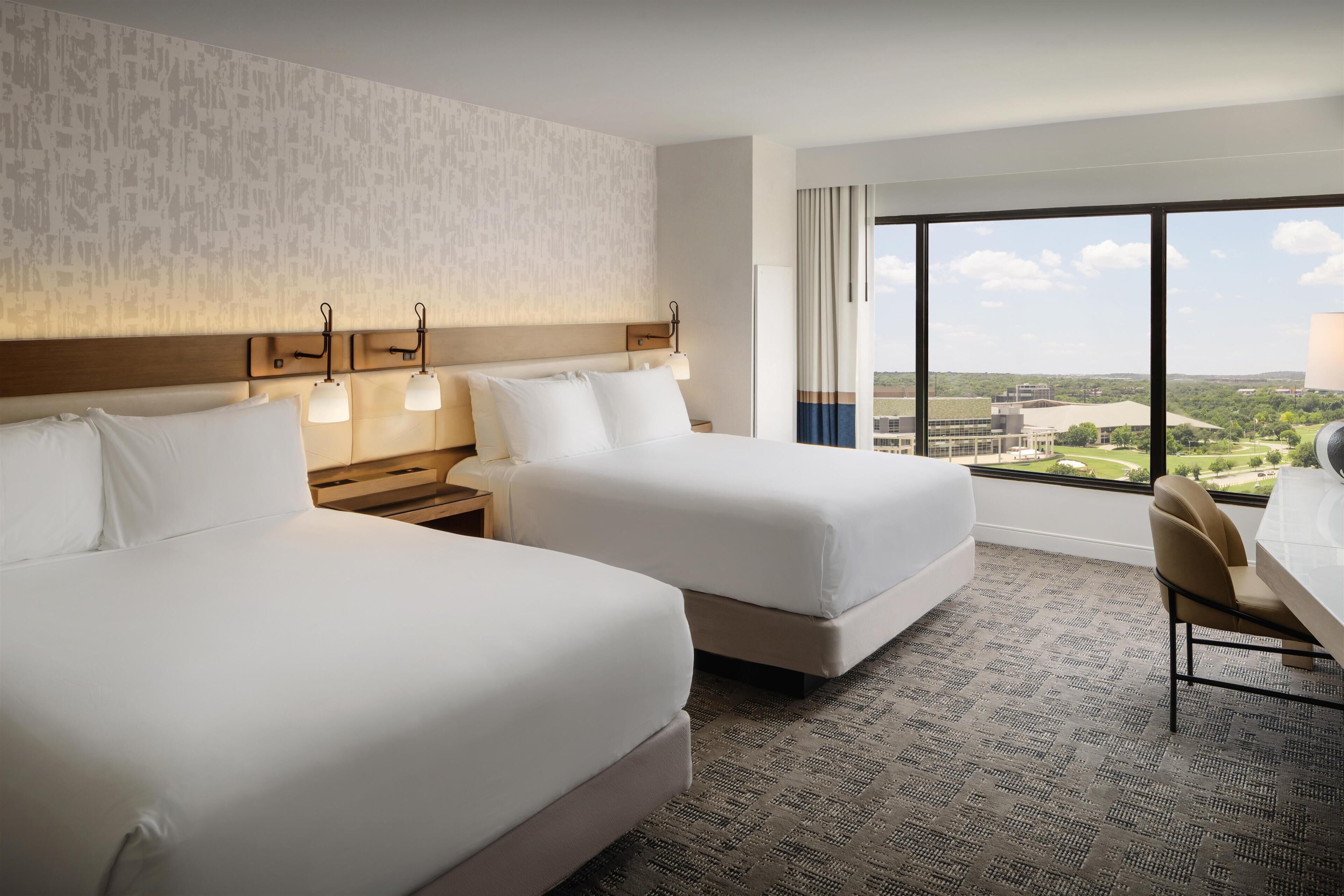 A hotel room with two beds, a desk, and a large window offering a view of the cityscape.