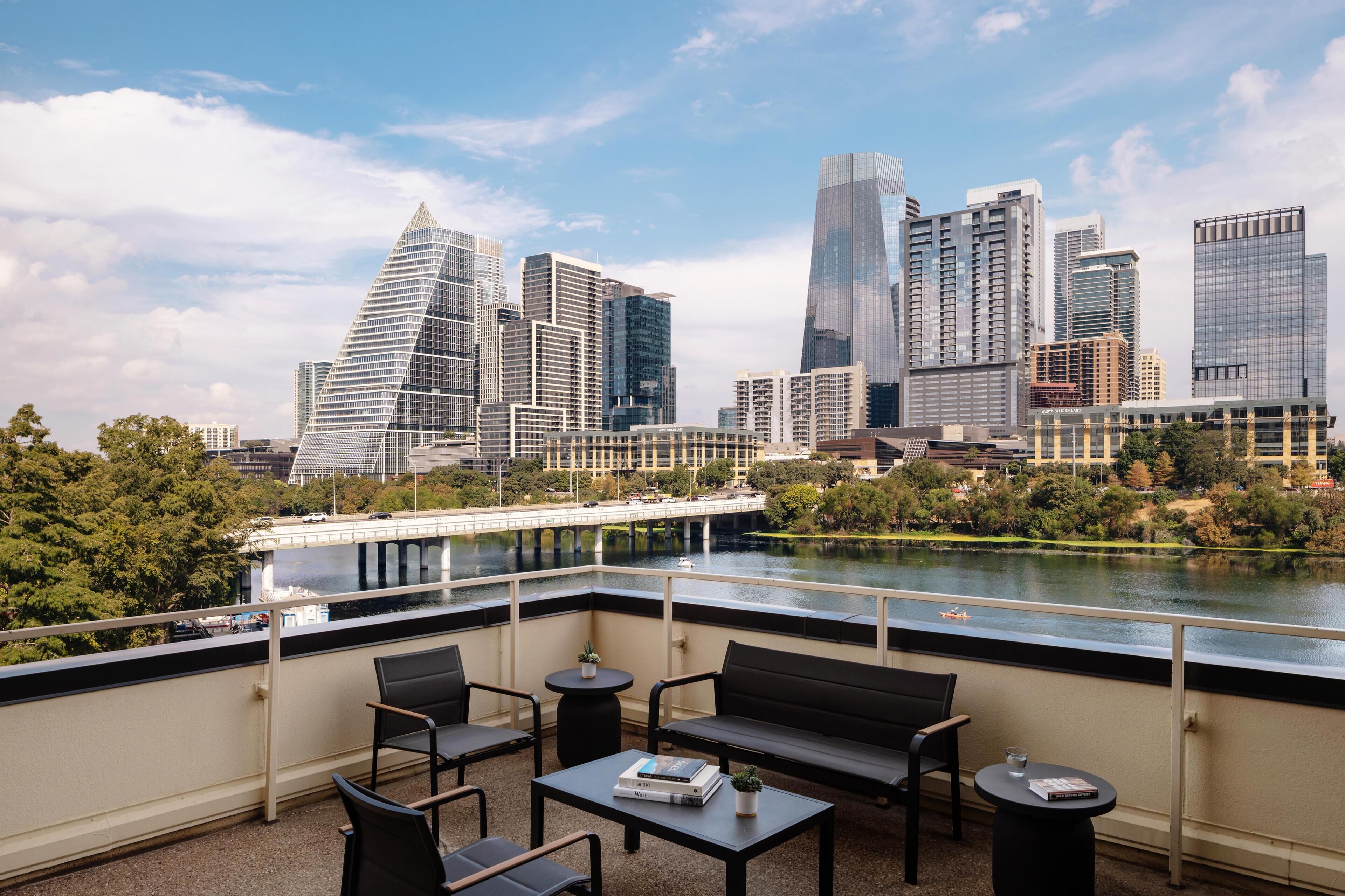A terrace with a view of a city skyline, including modern buildings and a river. There are chairs and tables for outdoor seating.