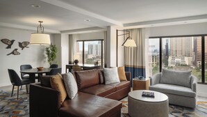 A modern hotel room with a brown leather sofa, grey armchair, round coffee table, dining area, and large windows offering city views. - Hyatt Regency Austin (Austin)