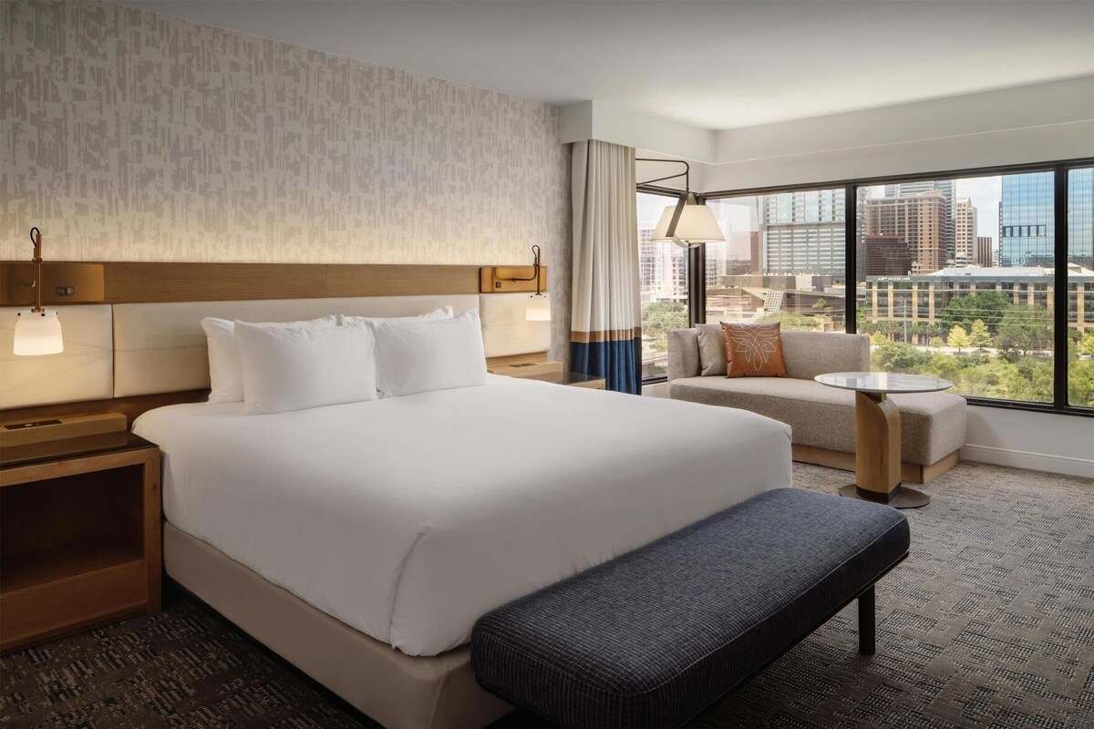 A hotel room with a large bed, a sofa, a small table, and a view of the city.