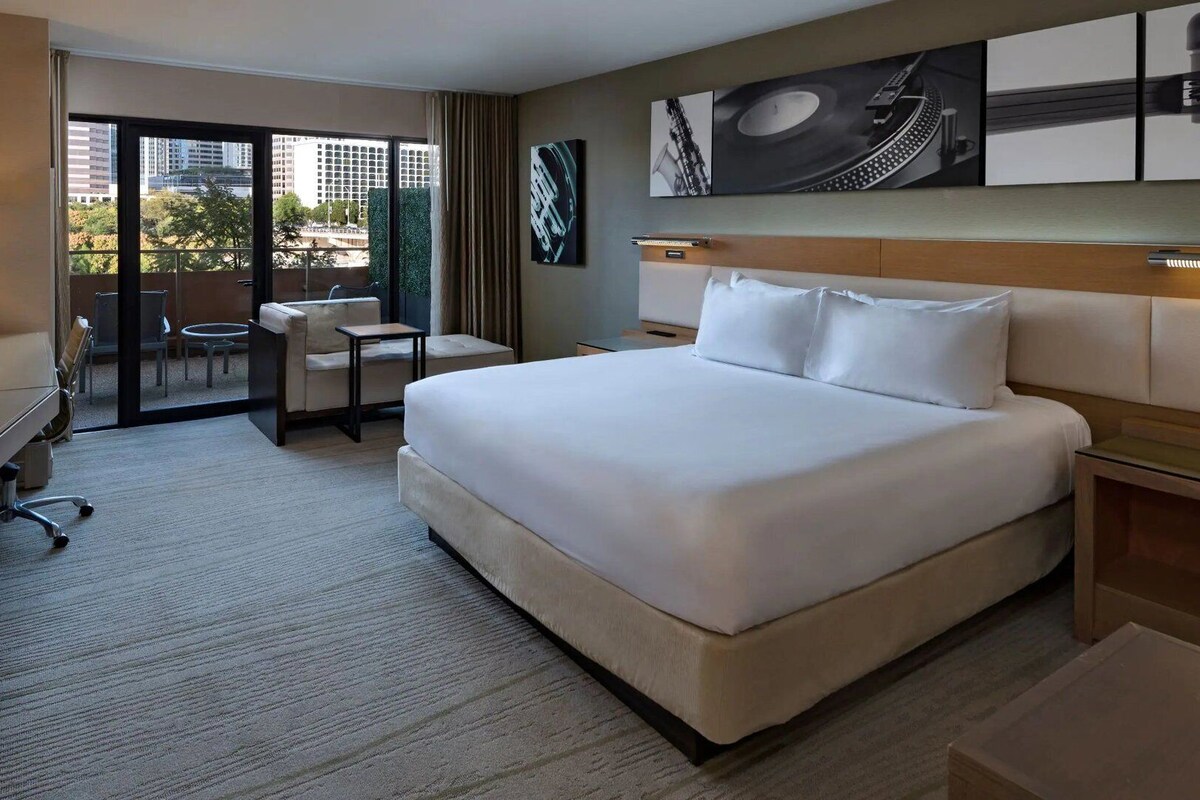 A hotel room with a large bed, a desk, a chair, and a view of the city through the window.