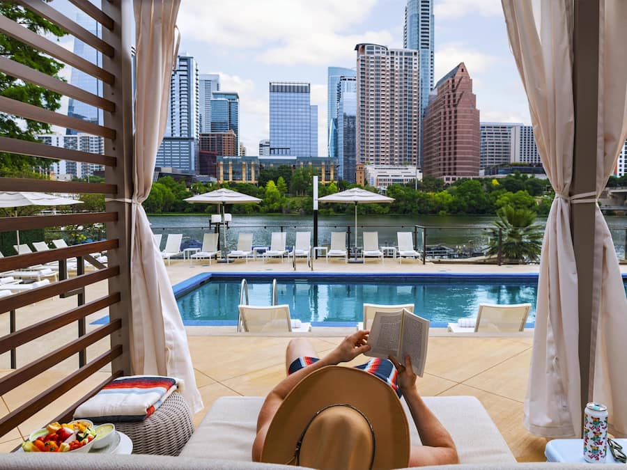 A person relaxing by a poolside with a city skyline view, lounge chairs, umbrellas, and a dining area with a fruit bowl.