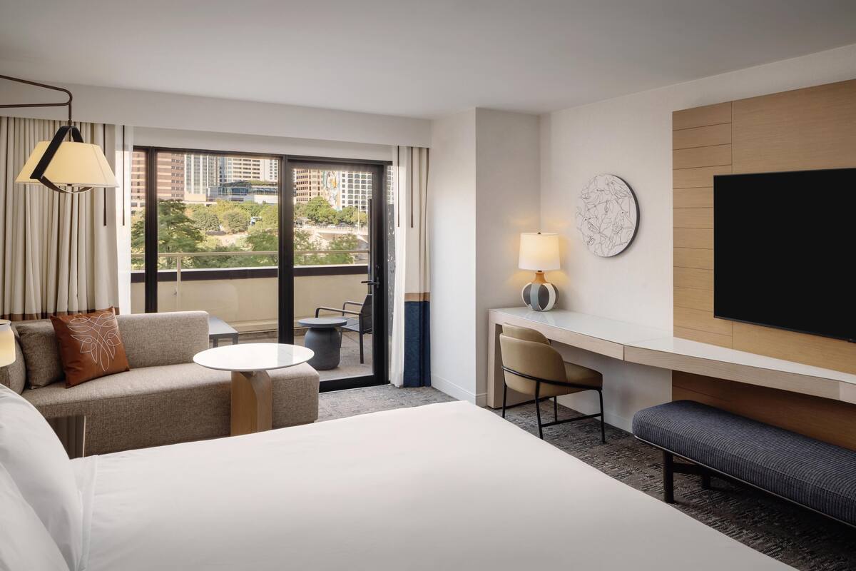 A hotel room with a bed, a sofa, a desk, a chair, a lamp, a TV, and a window with a view of buildings and trees.