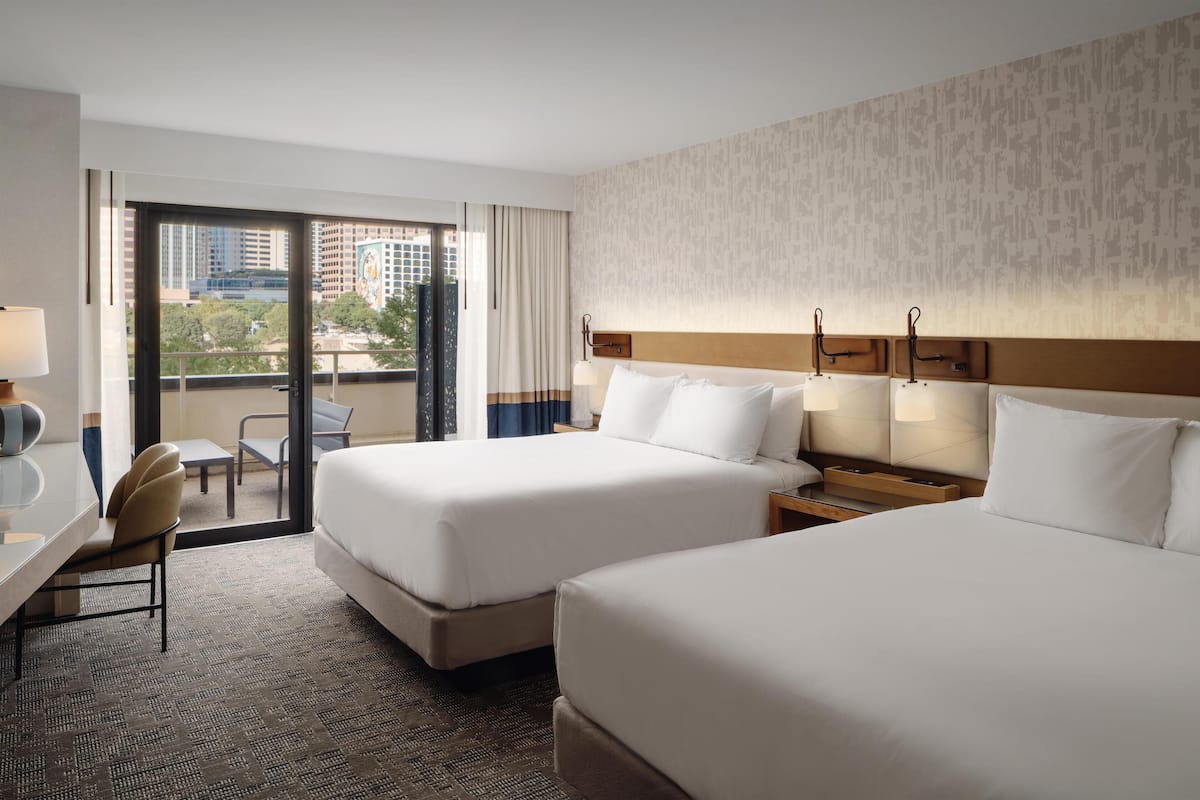 A hotel room with two beds, a desk, a chair, and a view of the city.