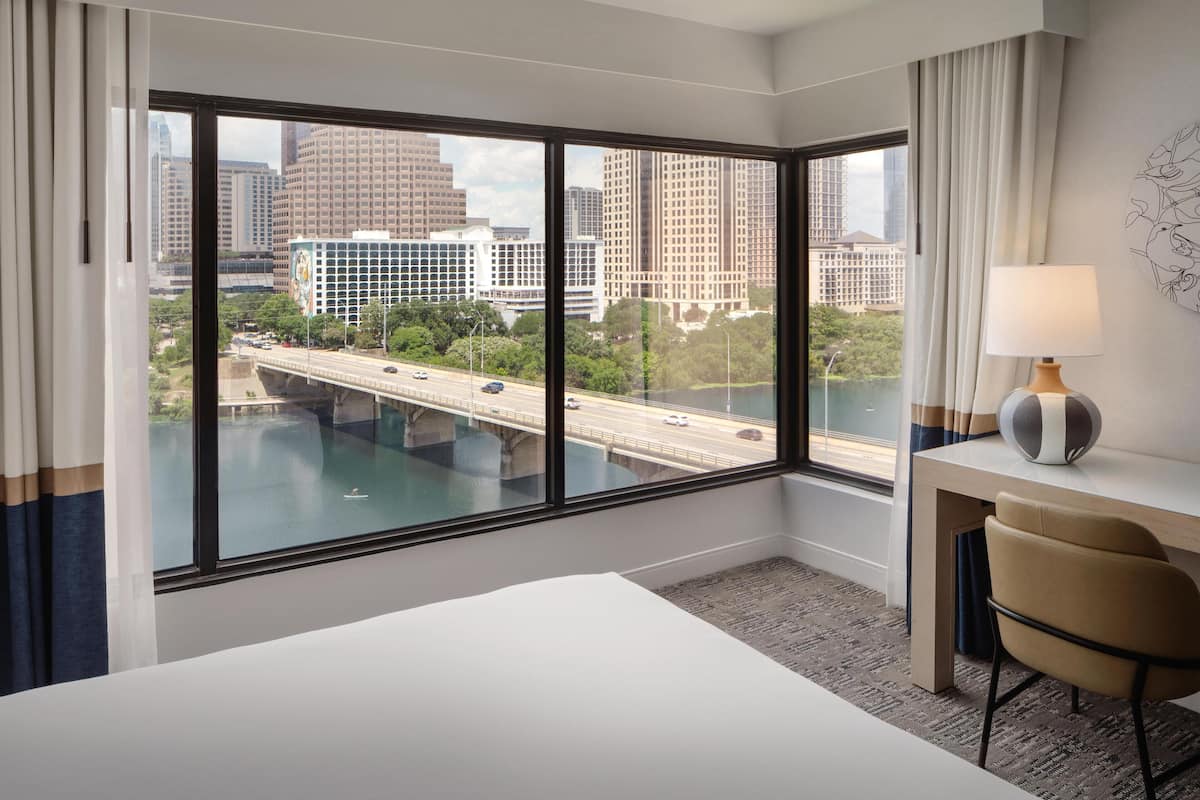 A hotel room with a large window, a bed, a desk with a lamp, and a chair.