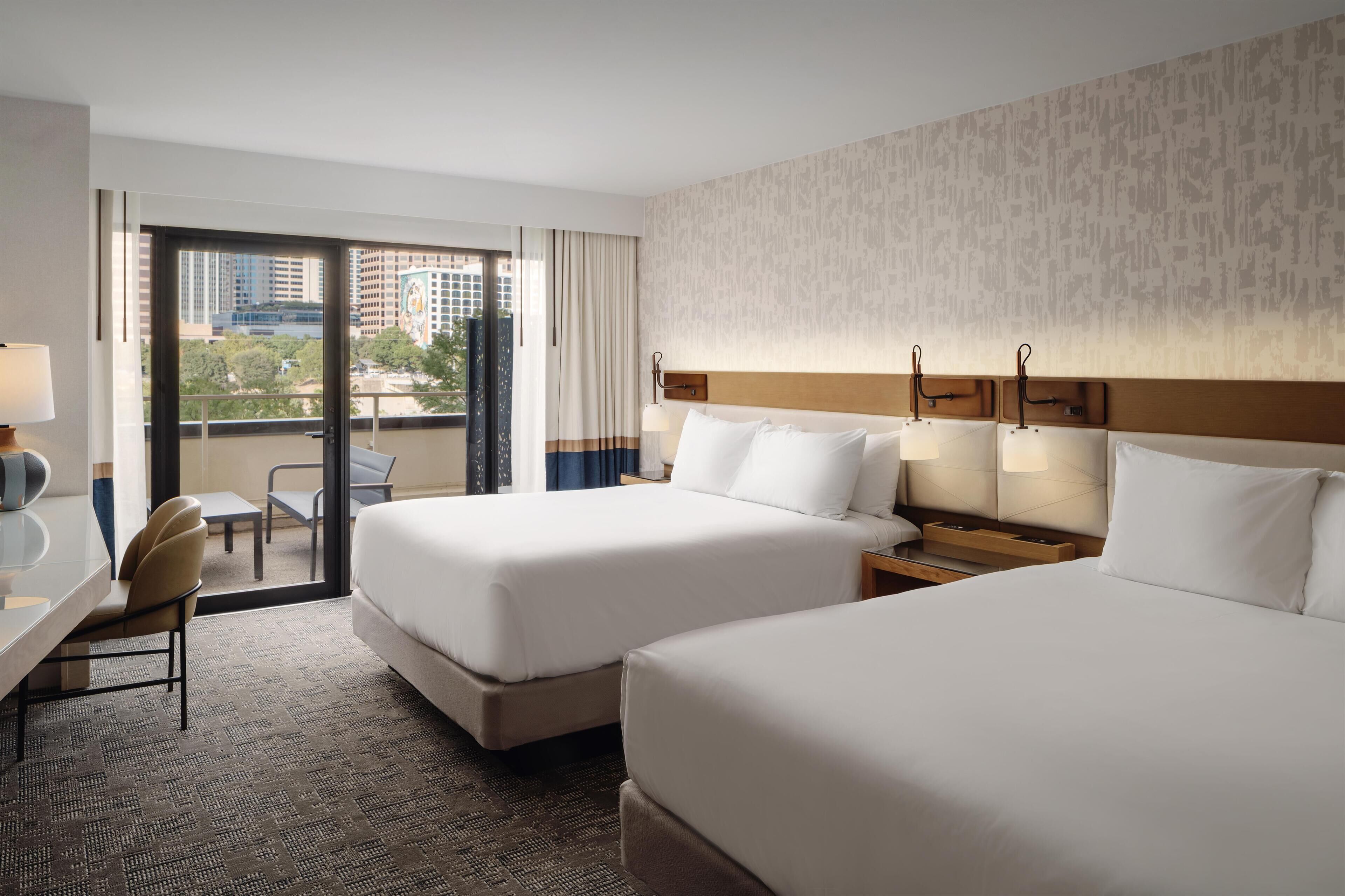 A hotel room with two beds, a desk, a chair, and a view of the cityscape through the window.