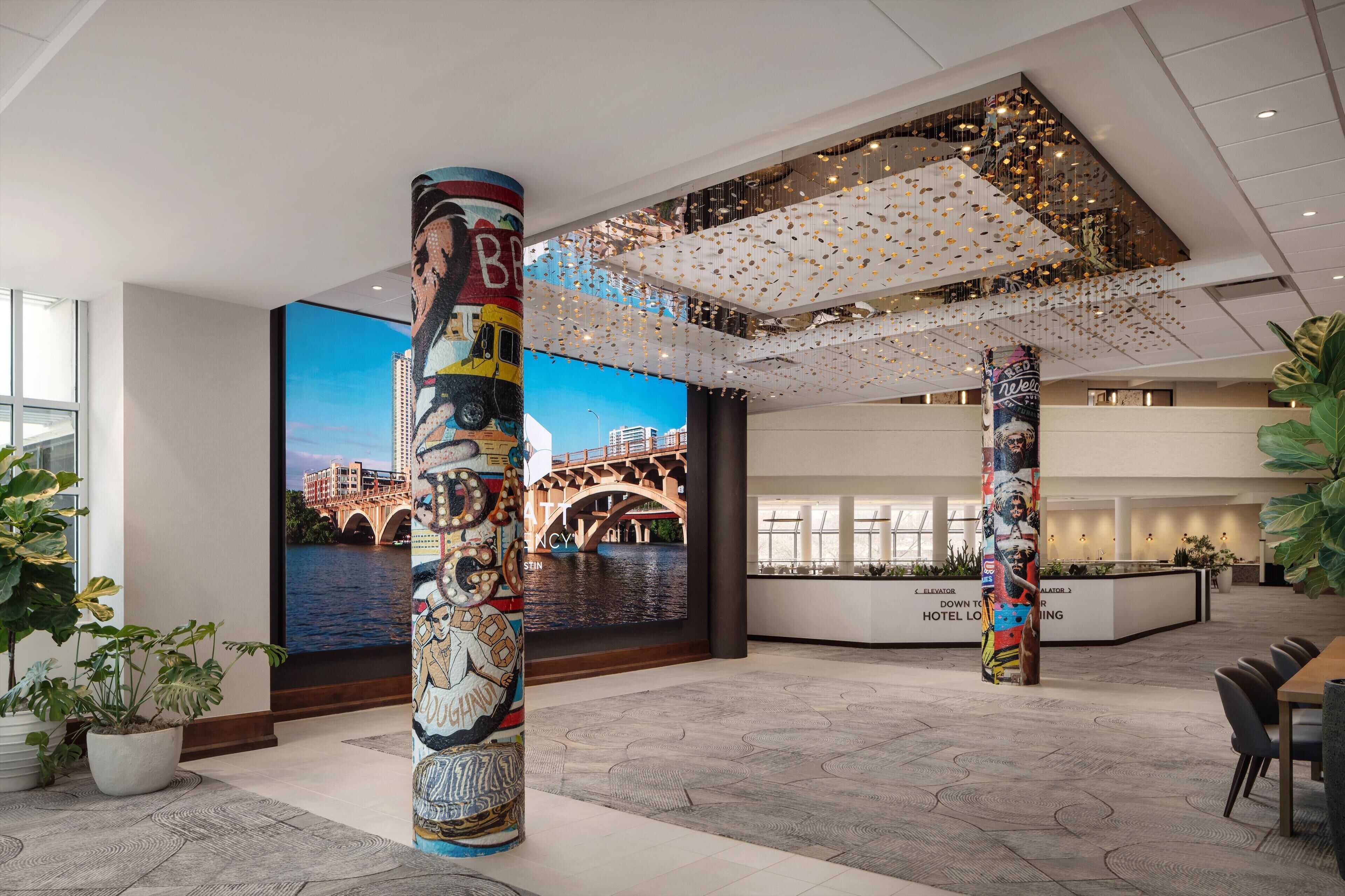 Un hall d’hôtel orné d’une grande fresque représentant un paysage urbain, avec un comptoir d’accueil et un design moderne au plafond, agrémenté d’ornements suspendus.