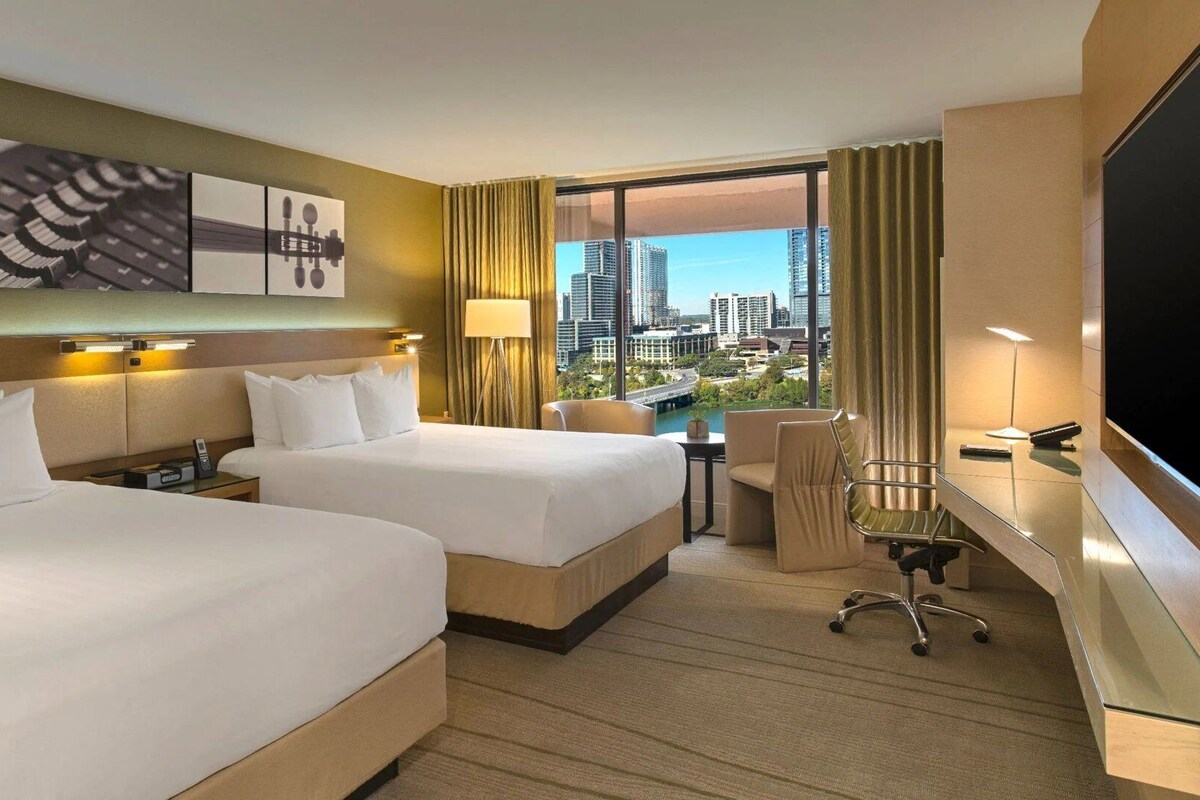 A hotel room with two beds, a desk, a chair, a TV, and a city view.