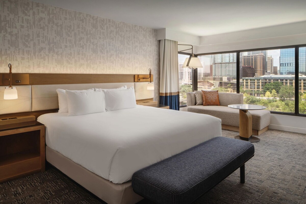 A hotel room with a large bed, a sofa, a small table, and a view of the cityscape.