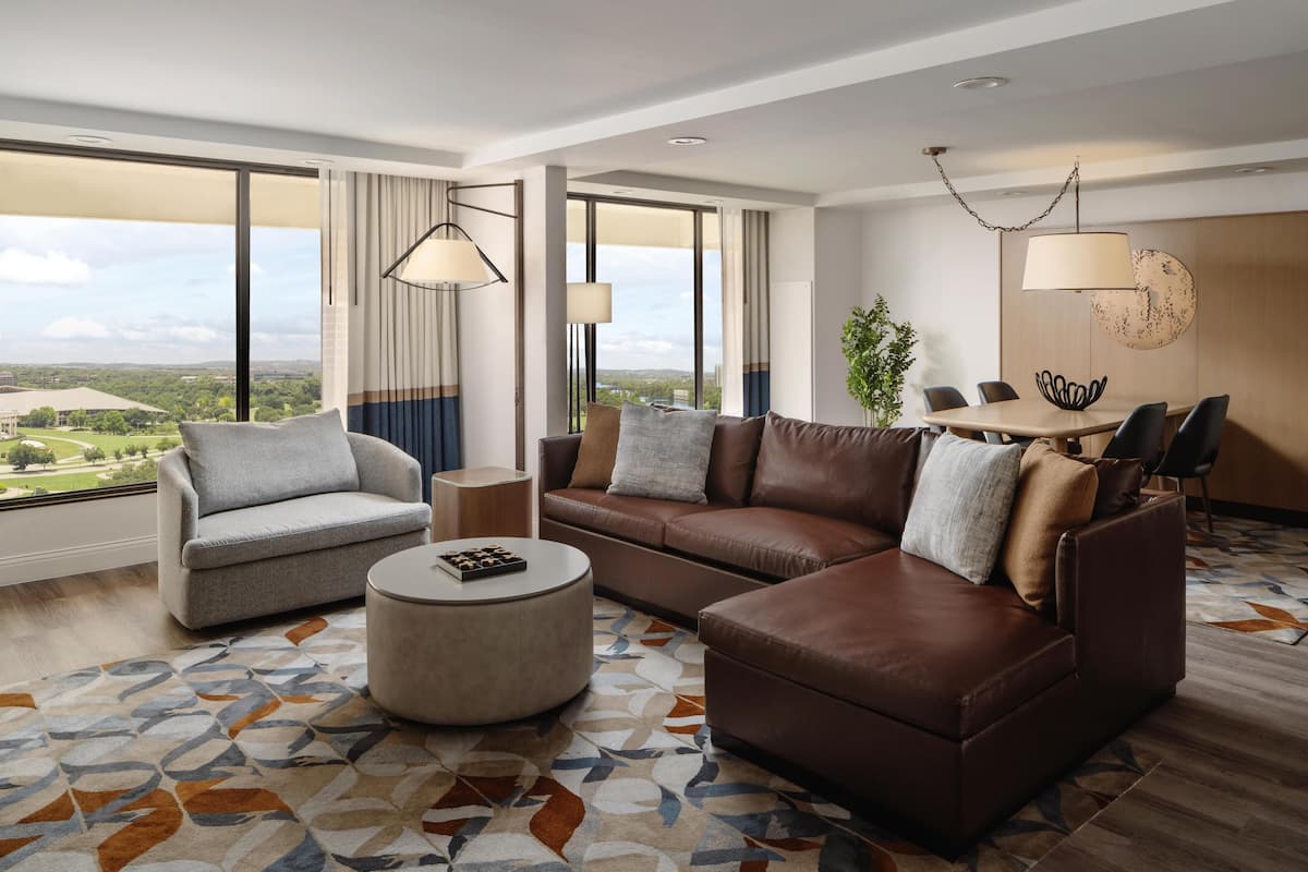 A modern hotel room with a large window, a brown leather sofa, a grey armchair, a round ottoman, and a dining table with chairs.