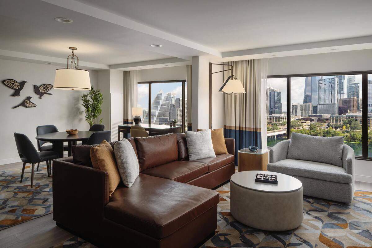 A modern hotel room with a leather sofa, a grey armchair, a round coffee table, and a dining area with a table and chairs.