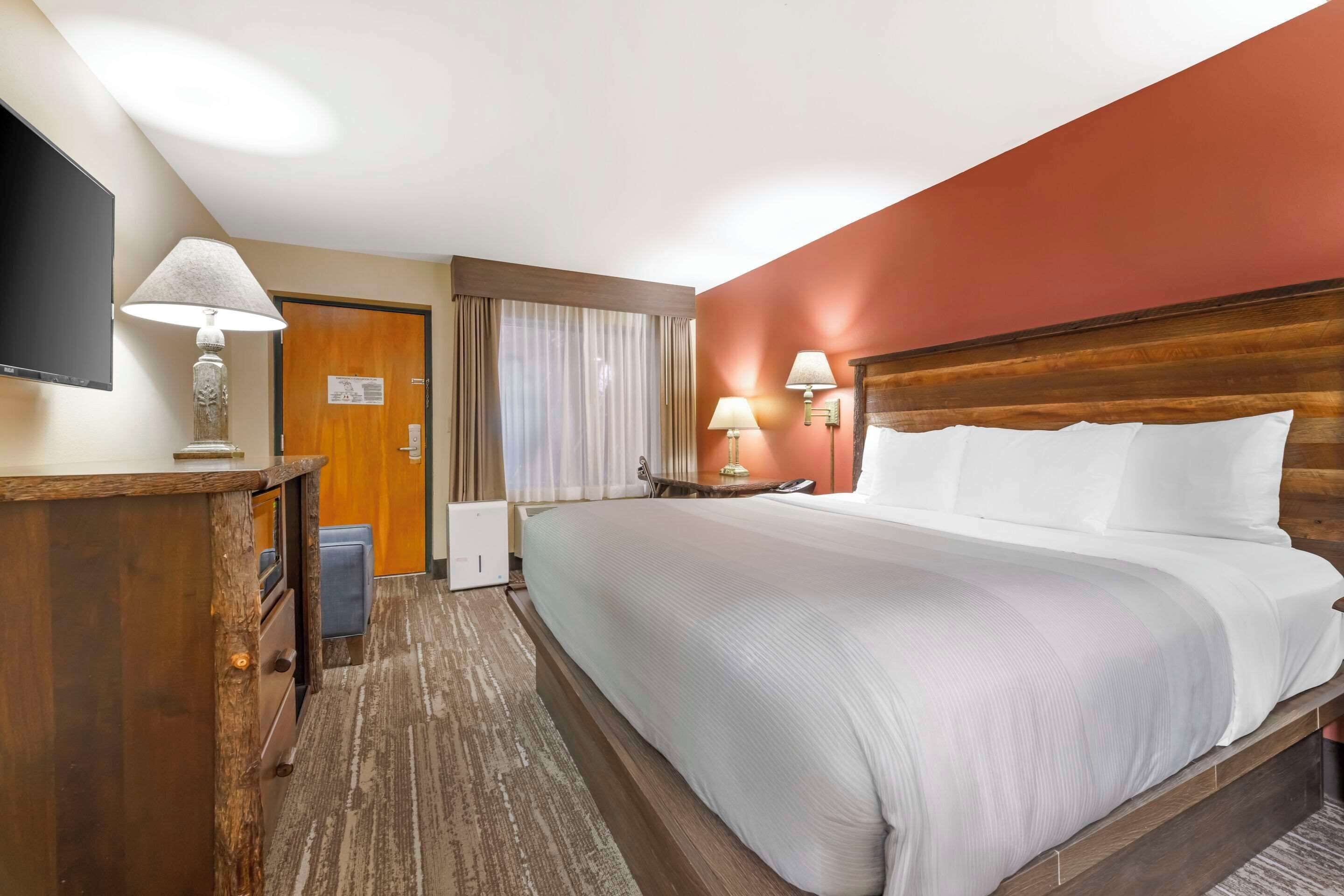 A hotel room with a large bed, a wooden headboard, a desk with a lamp, a flat-screen TV, and a window with curtains.