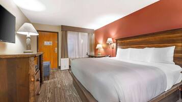 A hotel room with a large bed, a wooden headboard, a desk with a lamp, a flat-screen TV, and a window with curtains.