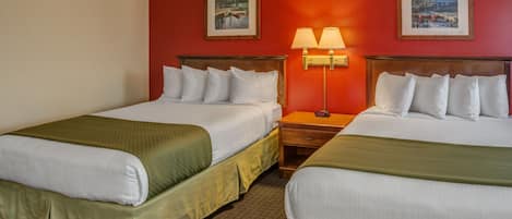 A hotel room with two beds, each with white linens and green bedspreads. There are two framed pictures on the walls, a nightstand with a lamp between the beds, and a carpeted floor.