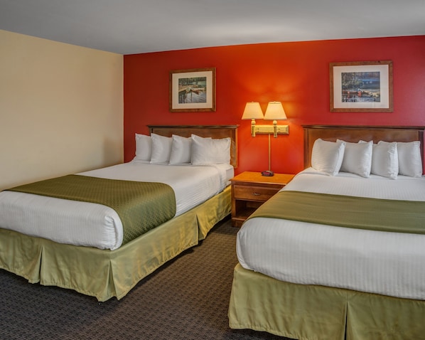 A hotel room with two beds, each with white linens and green bedspreads. There are two framed pictures on the walls, a nightstand with a lamp between the beds, and a carpeted floor.