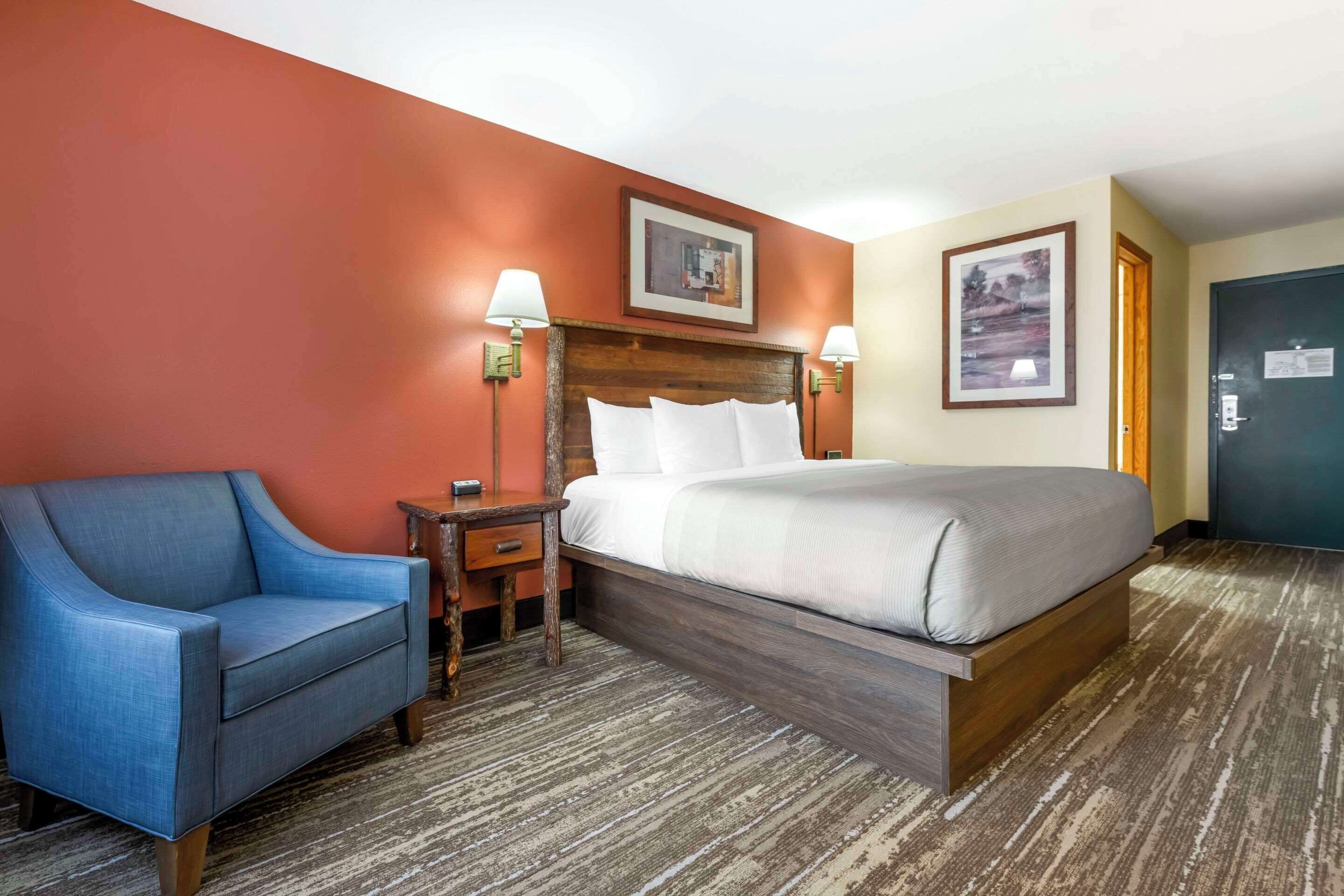 A hotel room with a bed, a blue armchair, a nightstand, two wall-mounted lamps, and two framed pictures.