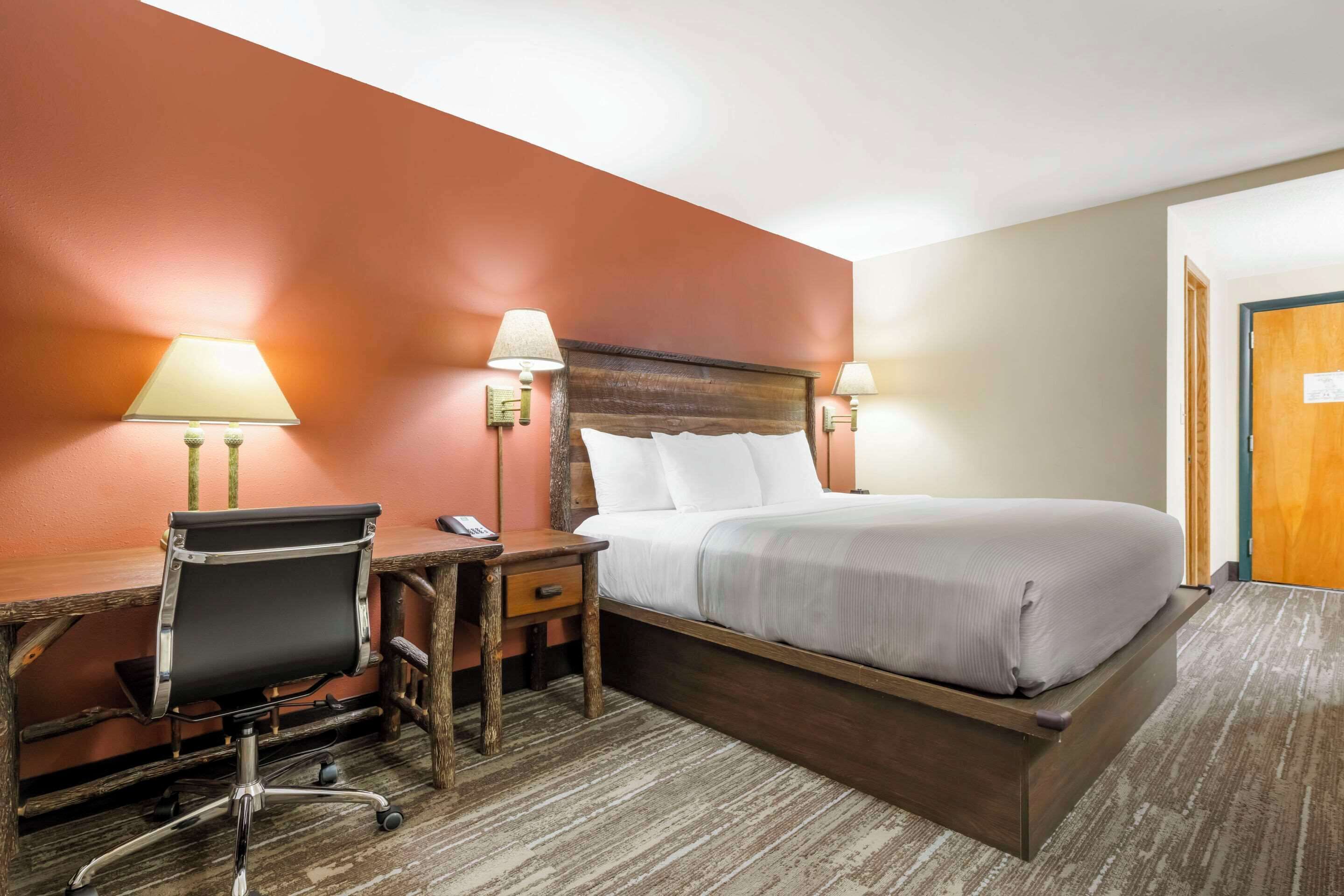 A hotel room with a wooden bed frame, a desk with a chair, two bedside lamps, and a carpeted floor.
