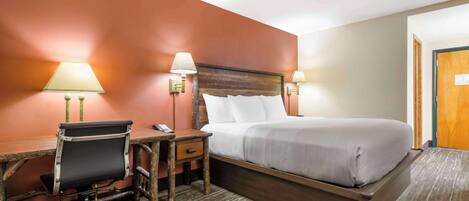 A hotel room with a wooden bed frame, a desk with a chair, two bedside lamps, and a carpeted floor.
