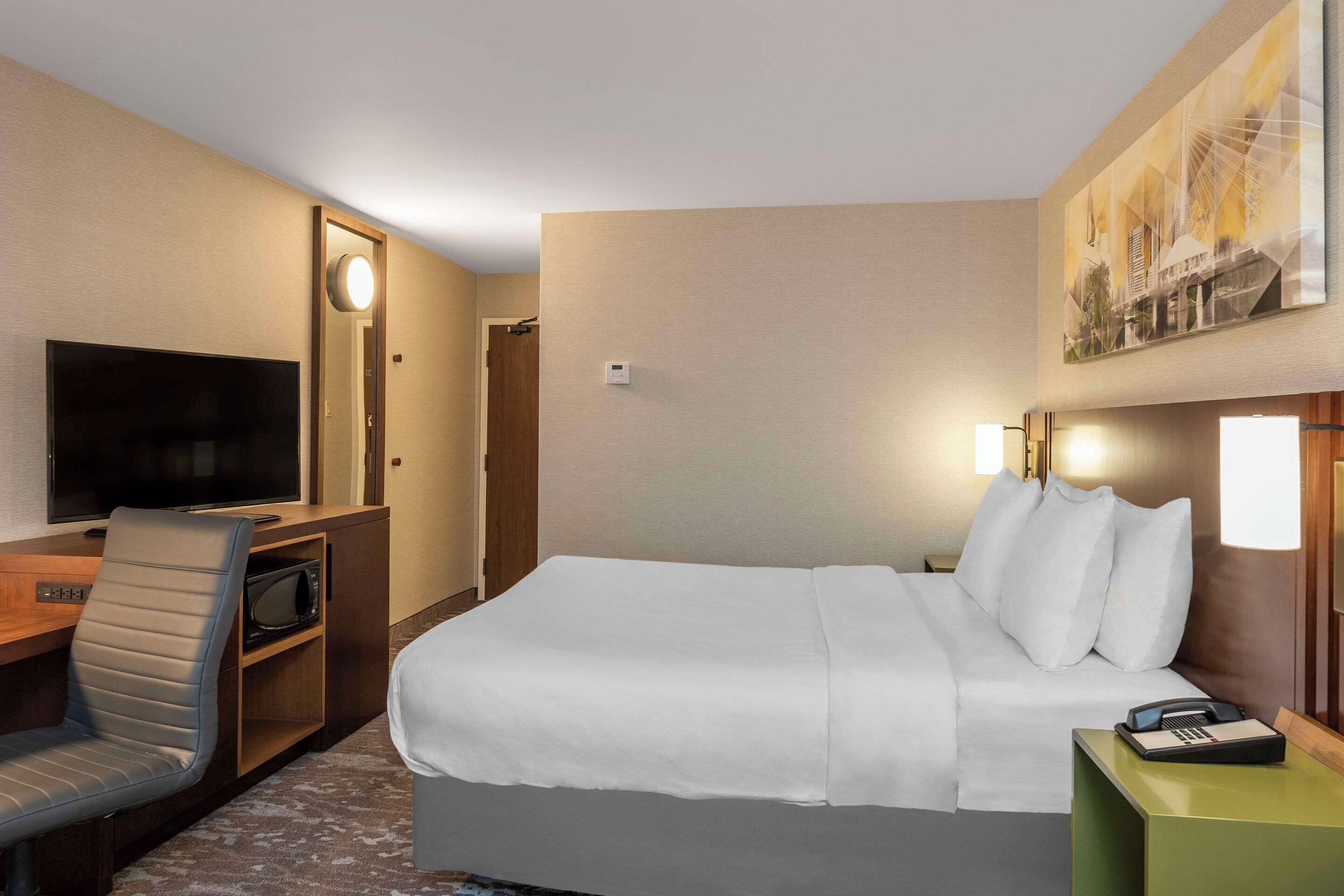 A hotel room with a bed, a desk with a chair, a television, and a painting on the wall.