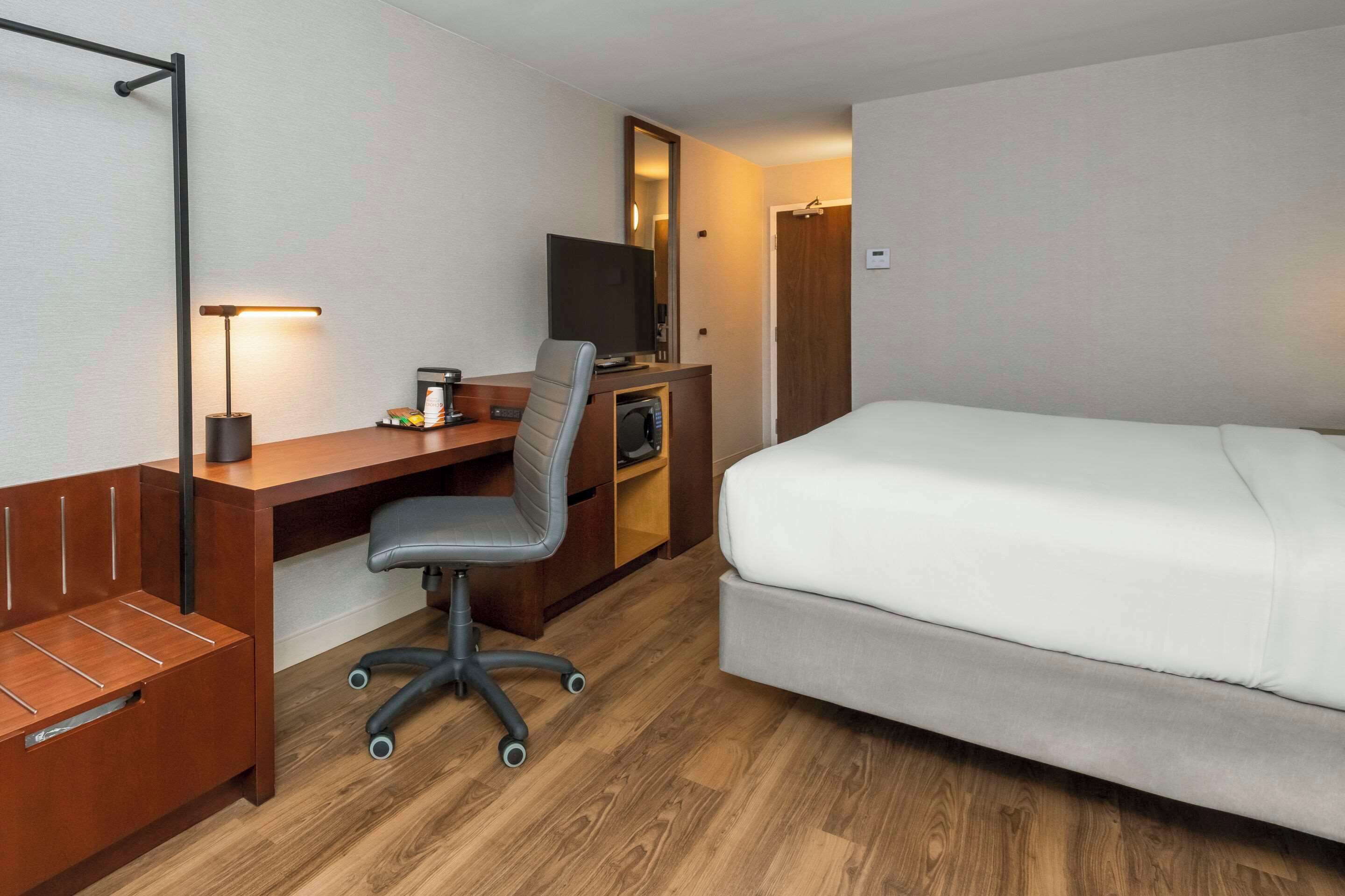 A hotel room with a bed, a desk with a chair, a TV, and a wooden bench.
