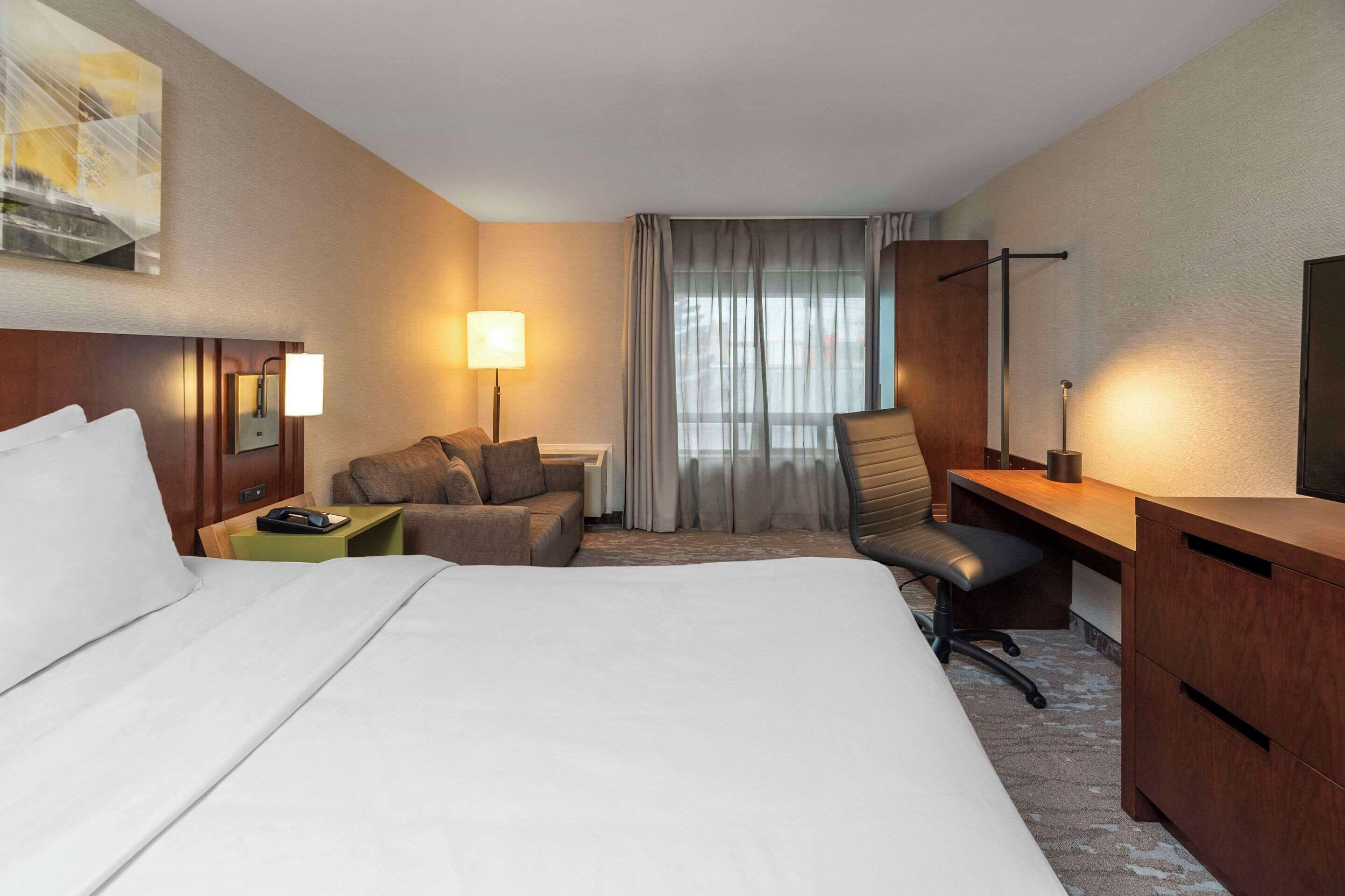 Ein Hotelzimmer mit einem großen Bett, einem Schreibtisch, einem Sessel, einem kleinen Tisch, einer Lampe, einem Fernseher und einem Fenster mit Vorhängen.