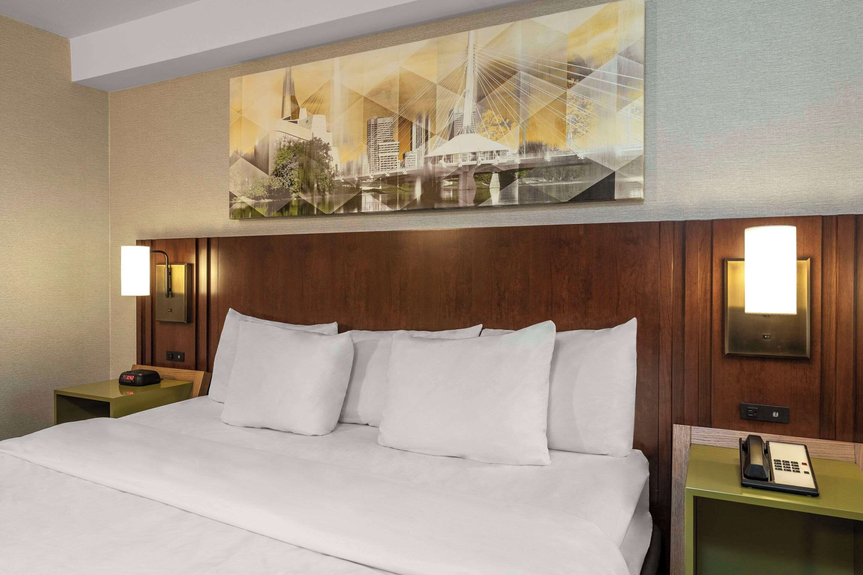 Habitación de hotel con una cama grande, cabecera de madera, dos mesitas de noche con lámparas y un cuadro con vista a la ciudad sobre la cama.