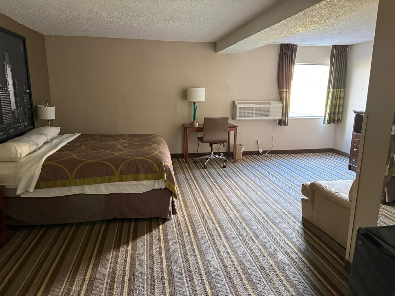 Ein Hotelzimmer mit Bett, Schreibtisch, Stuhl, Klimaanlage und einem Vorhangfenster.
