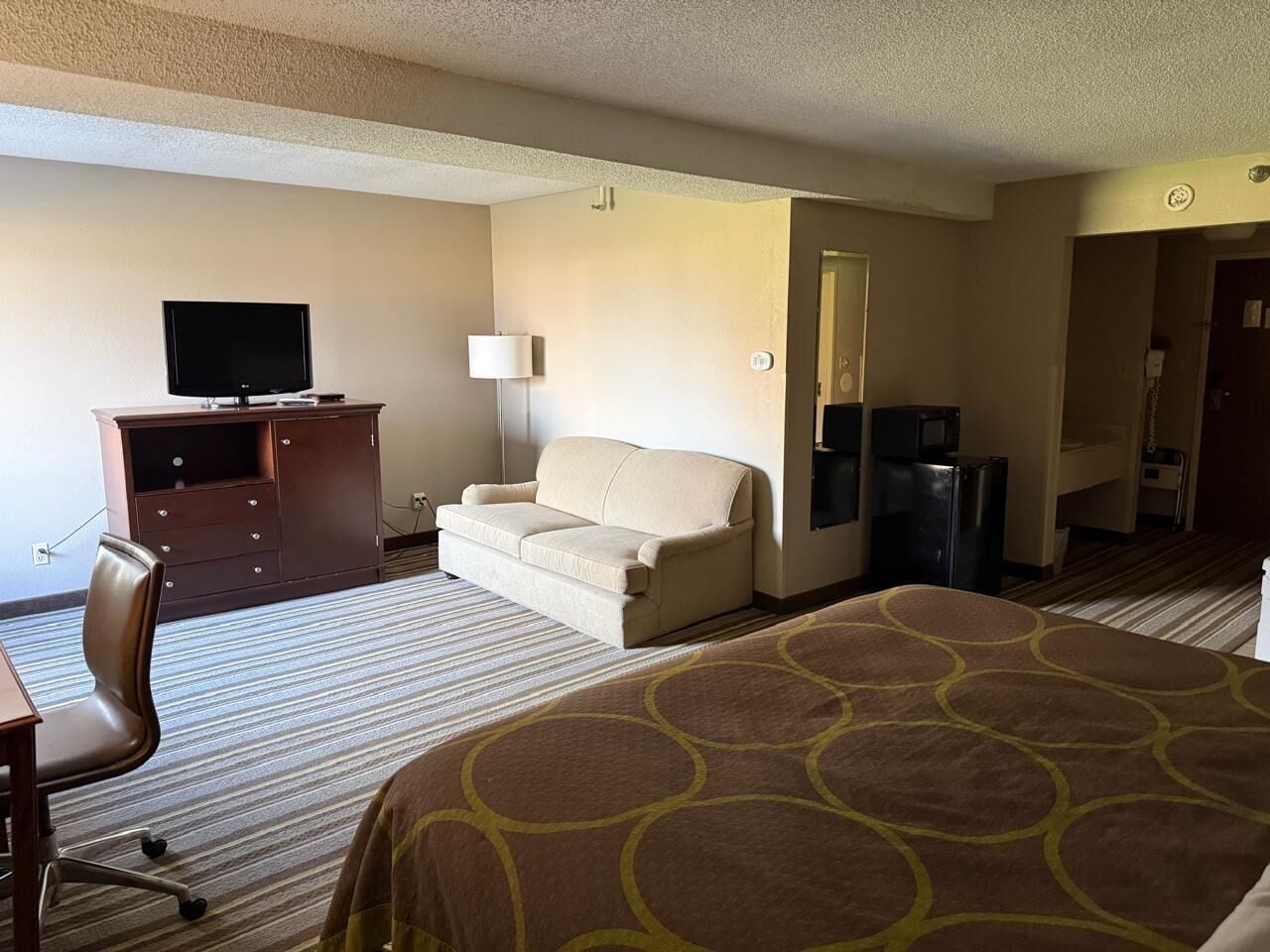 A hotel room with a bed, a desk, a chair, a sofa, a TV, and a refrigerator.