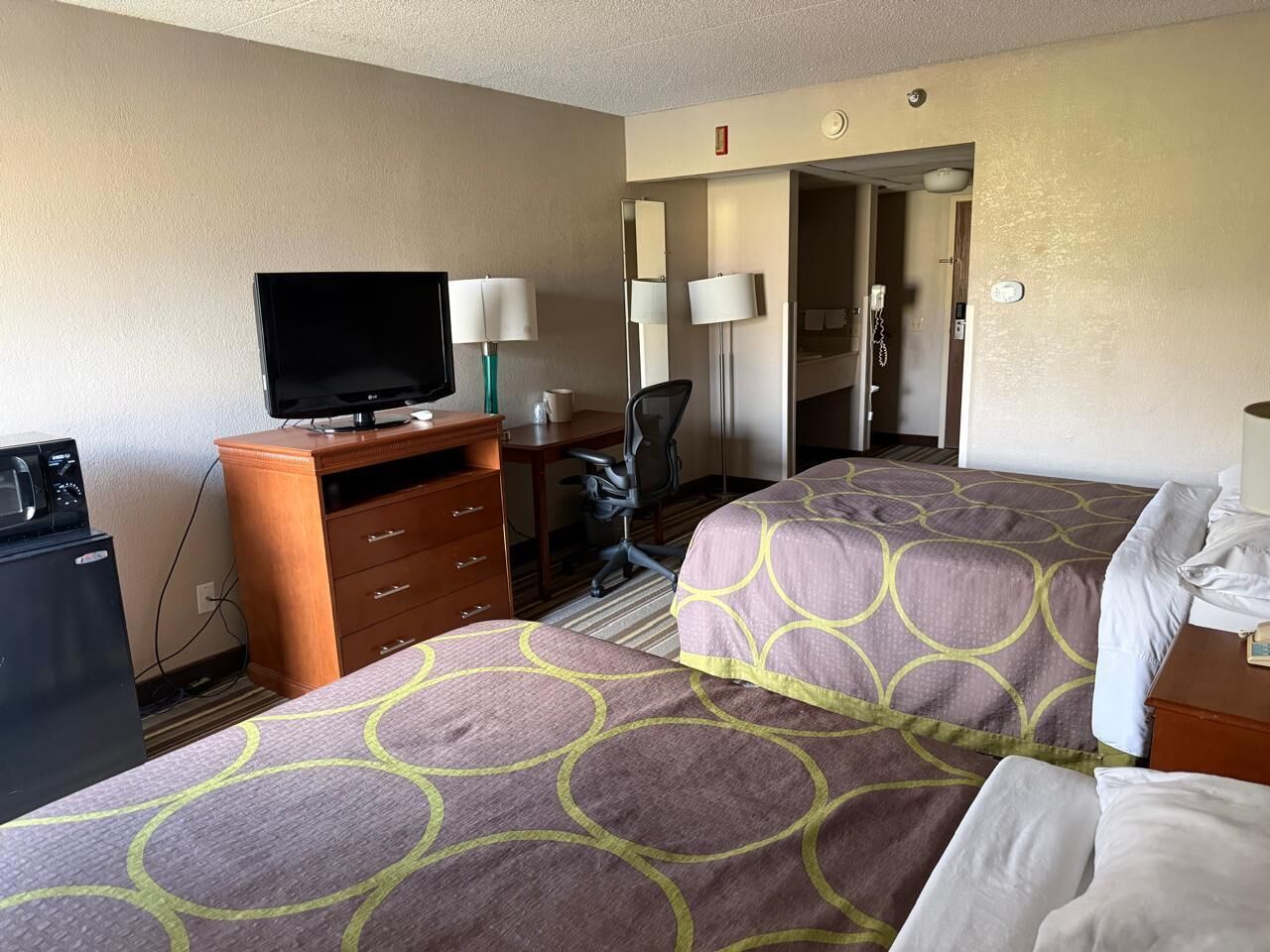 A hotel room with two beds, a desk with a television, a desk lamp, a chair, and a minibar.
