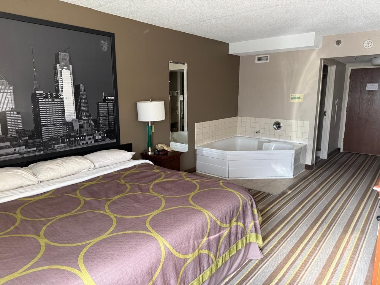 A hotel room with a bed, a cityscape wall, a bathroom with a bathtub, and a striped carpet.