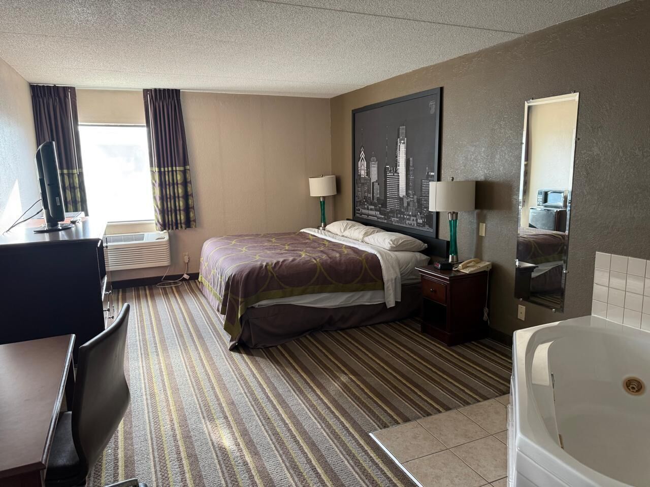 A hotel room with a bed, desk, chair, lamp, and a large window with curtains.