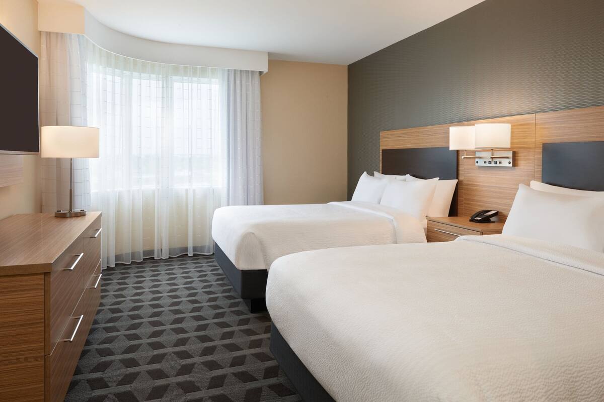 A hotel room with two beds, a desk, a lamp, and a window with sheer curtains.