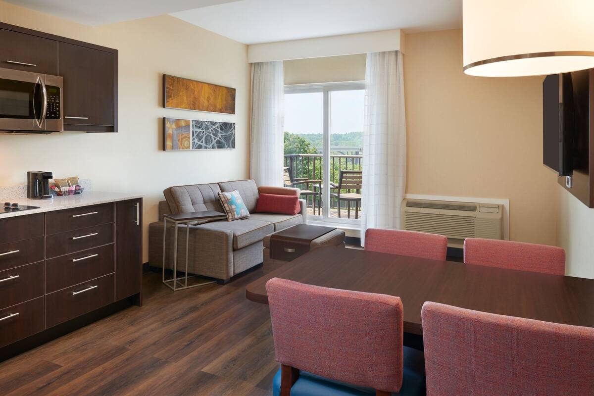 A modern hotel room with a dining area, a sofa, a flat-screen TV, and a balcony with a view.