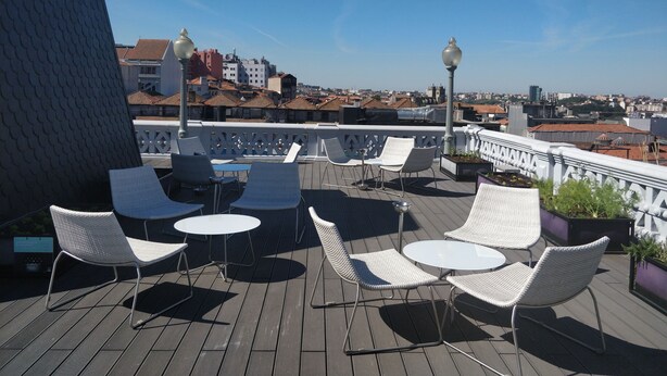 A rooftop terrace with white wicker chairs and tables, a railing, and a view of a cityscape. - Grande Hotel do Porto (Porto)