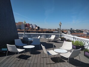 A rooftop terrace with white wicker chairs and tables, a railing, and a view of a cityscape. - Grande Hotel do Porto (Porto)