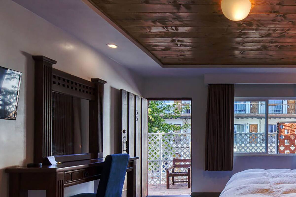 Habitación de hotel con techo de madera, cama con ropa blanca, escritorio con silla, ventana con cortinas y mesita de noche con lámpara.