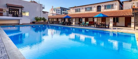 A swimming pool with lounge chairs and umbrellas, surrounded by buildings with balconies and windows.
