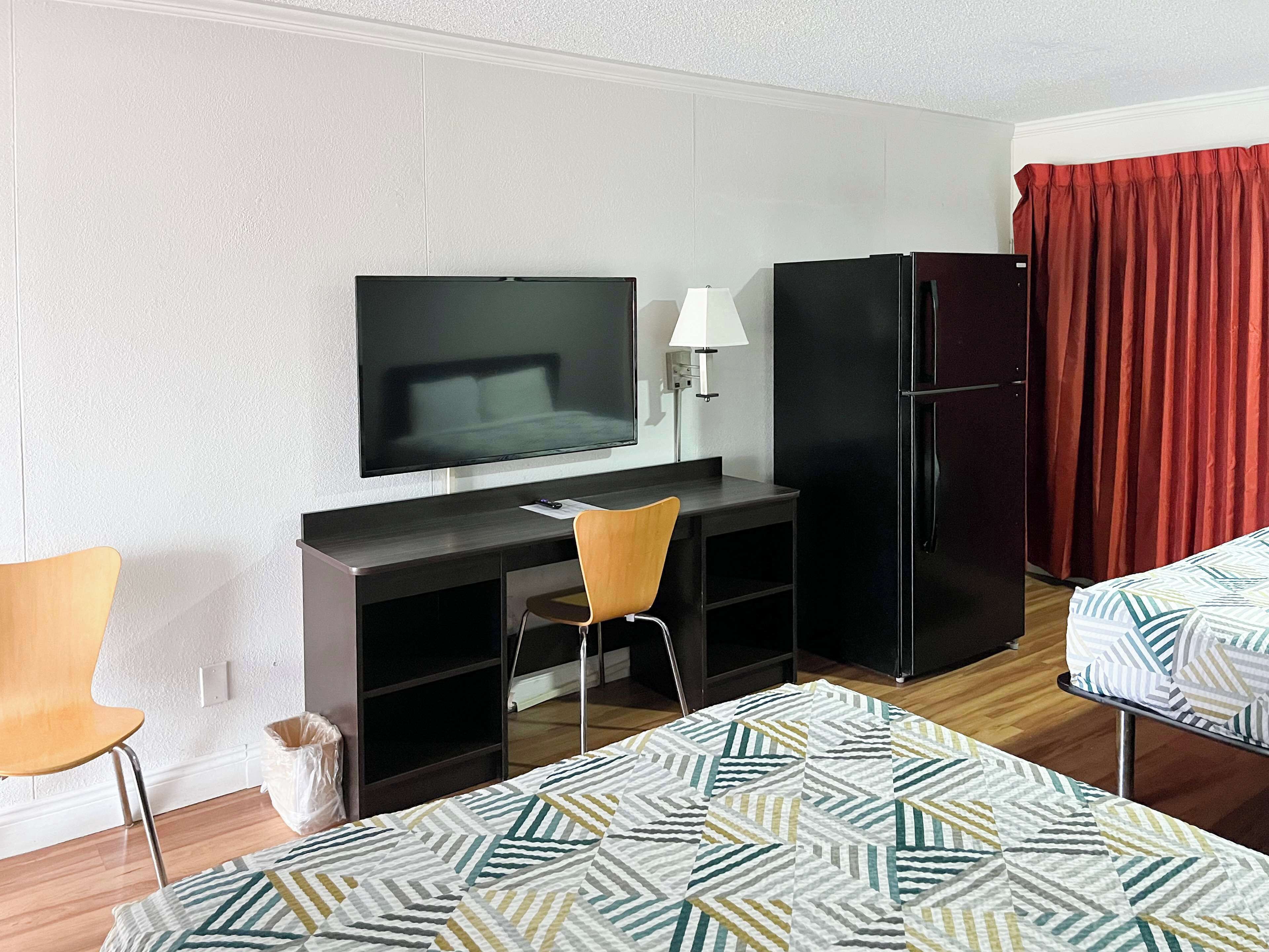 A compact hotel room with a bed, a desk with a television, a refrigerator, and a chair.