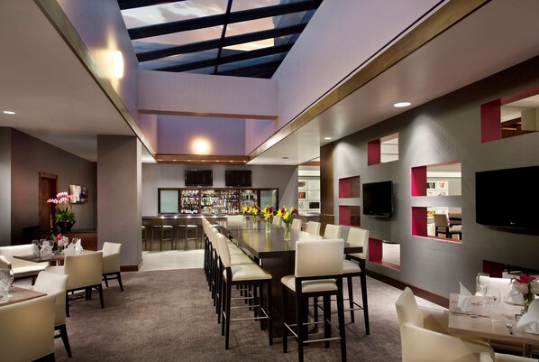 A modern dining area with a long table, multiple chairs, a bar area, and a television mounted on the wall.